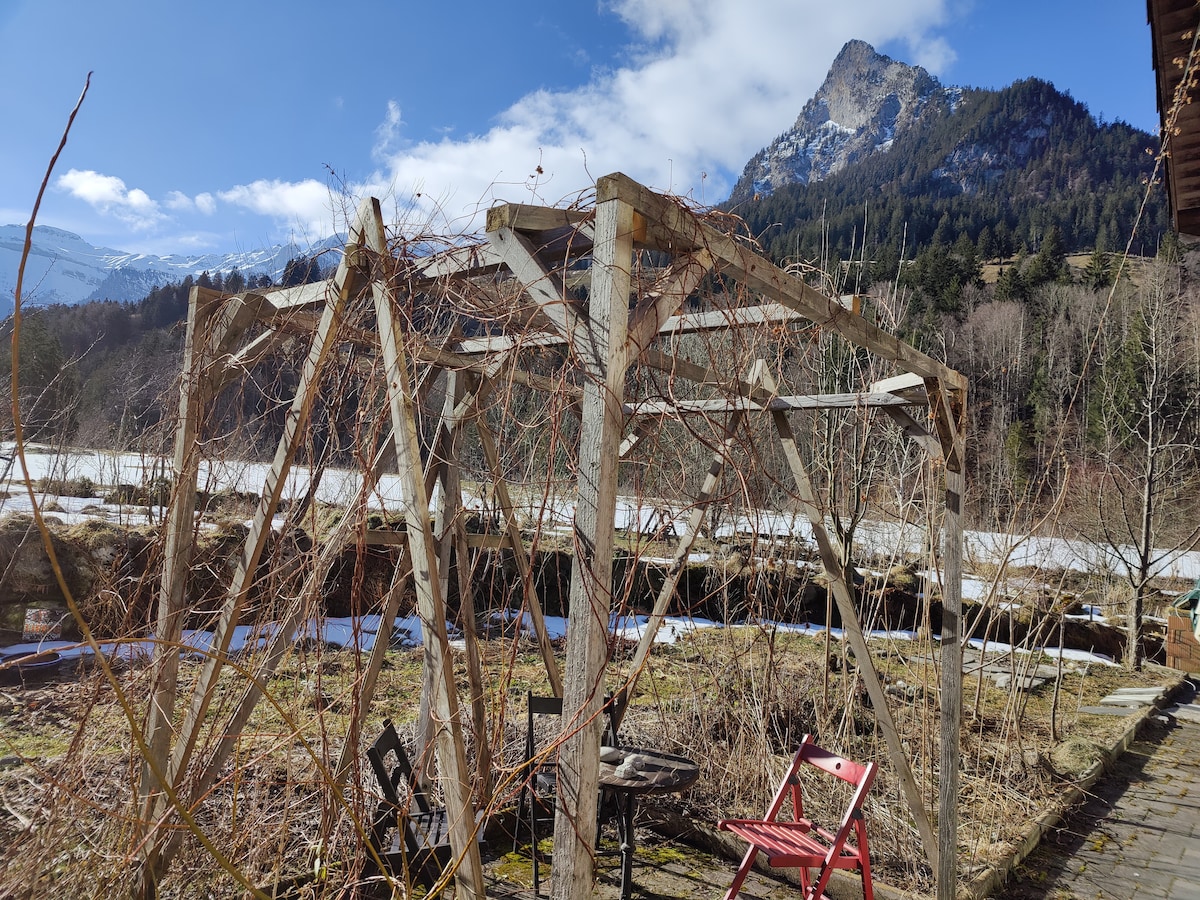 An open outdoor space is visible, featuring a wooden trellis with climbing plants. Two chairs, one red and one dark, are positioned nearby. The backdrop displays a mountain landscape under a partly cloudy sky, creating a natural setting for relaxation.
