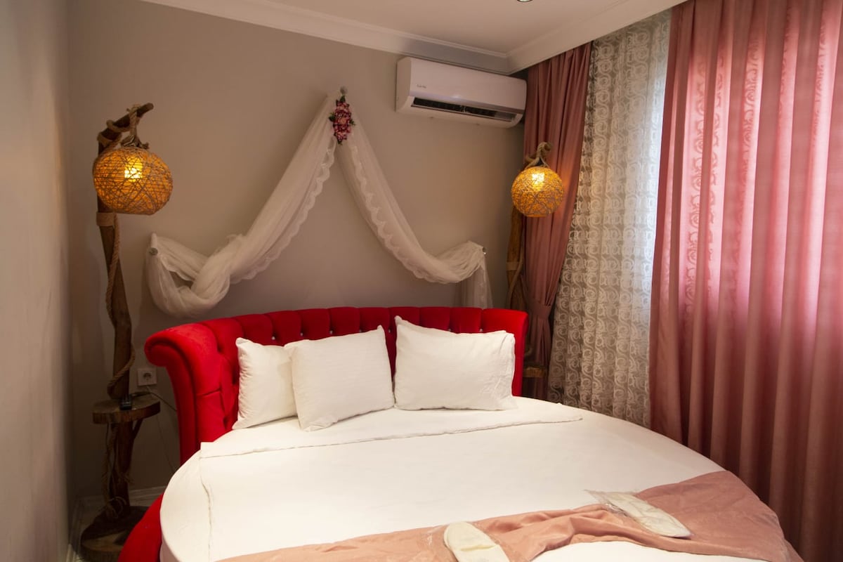 A round bed with a plush red headboard is positioned against a wall, complemented by decorative pillows. Elegant lighting fixtures hang above, and sheer curtains softly filter natural light. A light-colored drape is arranged on the wall, adding texture to the space.
