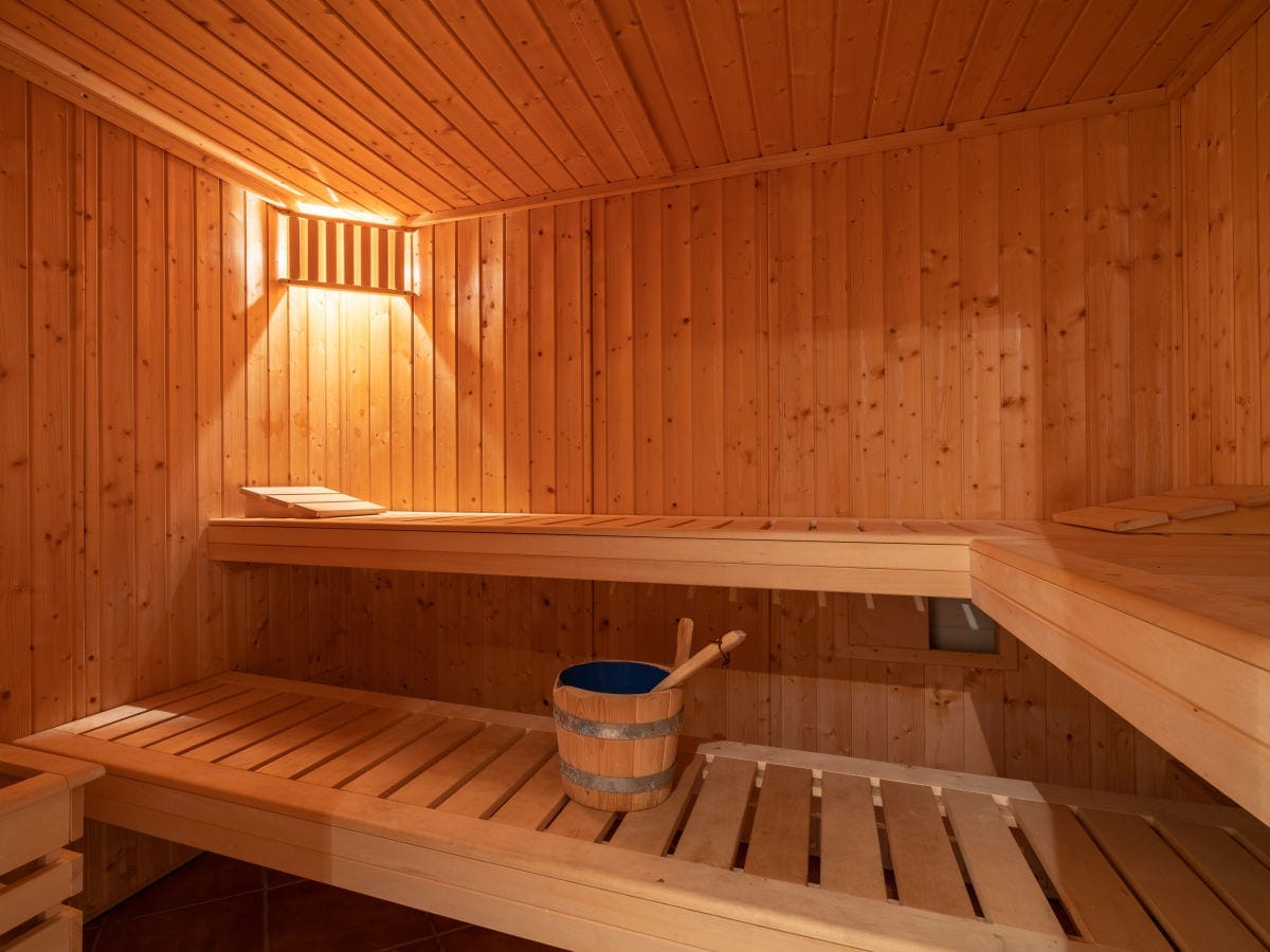 A wooden sauna interior features warm, natural wood paneling. The space includes slatted seating areas on two levels, with a blue bucket and wooden scoops positioned on a shelf. Soft light is cast overhead, enhancing the serene environment.