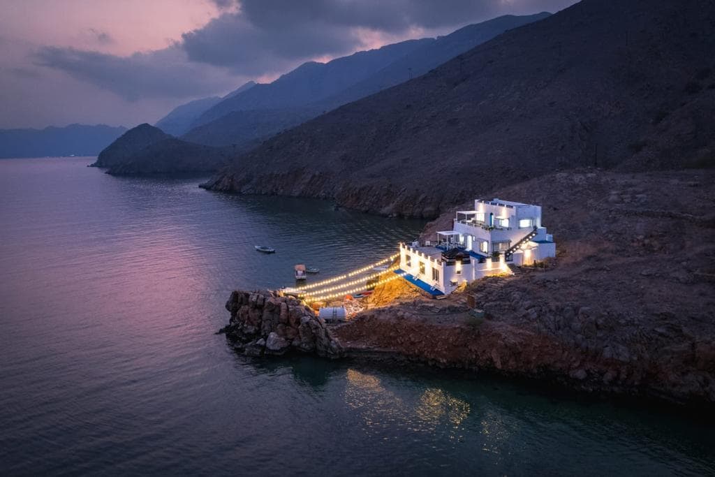 A villa is positioned on a rocky shoreline, surrounded by calm water reflecting evening light. Soft illumination highlights the structure, enhancing its presence against the backdrop of mountainous terrain. Gentle waves lap at the rocks, emphasizing the serene atmosphere of the secluded location.