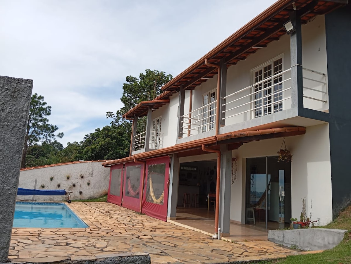 The two-story property features a combination of light and dark exterior walls, with large windows and a balcony on the upper level. A stone patio surrounds a private swimming pool, while vibrant greenery is visible in the background.