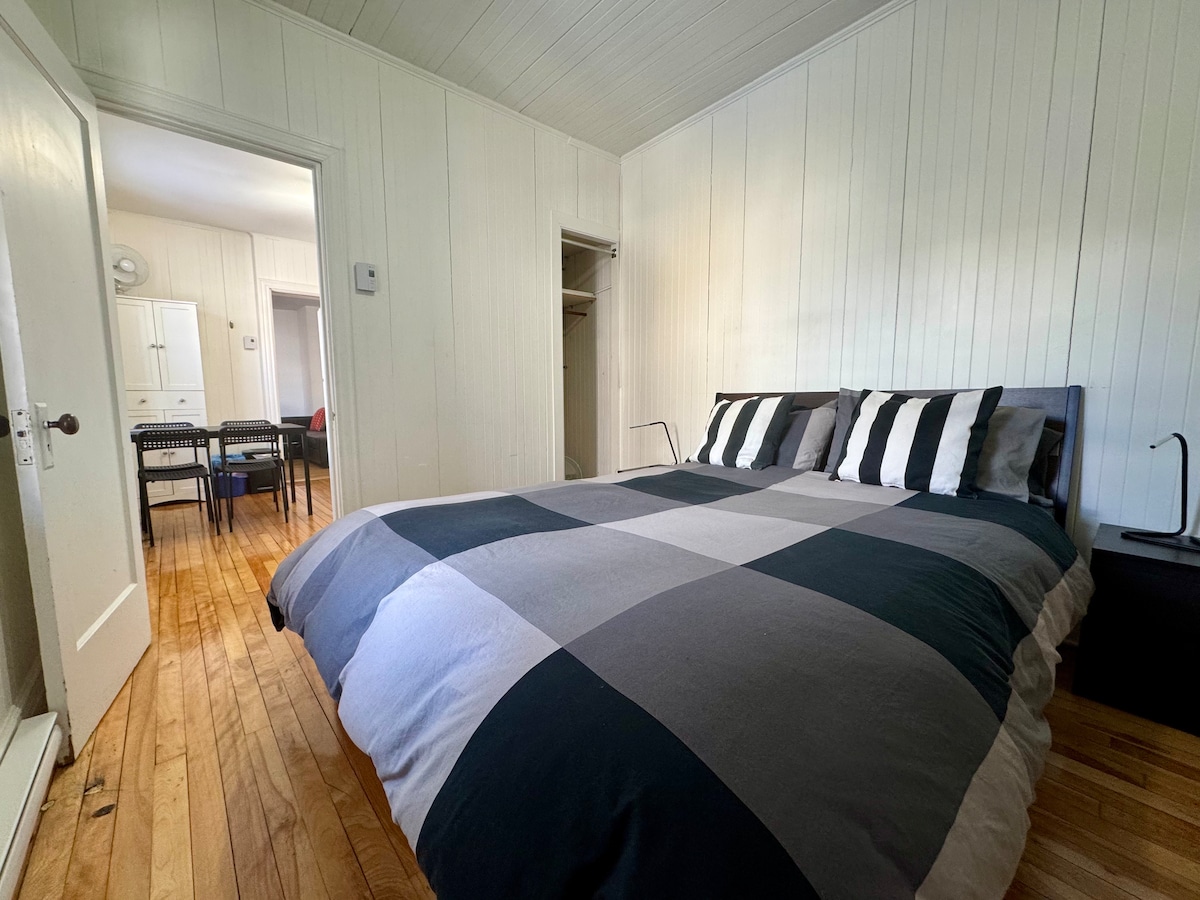 The bedroom features a spacious bed topped with a checkered duvet in shades of gray and black. Light hardwood flooring enhances the room's brightness. A nightstand with a lamp is positioned beside the bed, while a closet and doorway leading to an adjacent area are visible.
