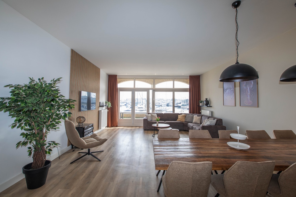 A spacious living area is presented with large windows allowing ample natural light and views of the harbor. A cozy seating arrangement, featuring a sectional sofa and armchair, is accompanied by a dining table with several chairs. A potted plant enhances the inviting atmosphere.