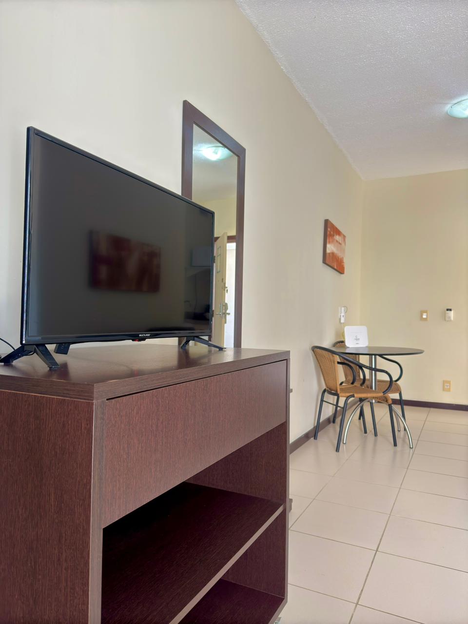 A modern living area features a dark wood TV stand with a large flat-screen television, complementing the light-colored walls. A round table with four chairs occupies the corner, providing a space for dining or work. A mirror reflects the room's natural light.