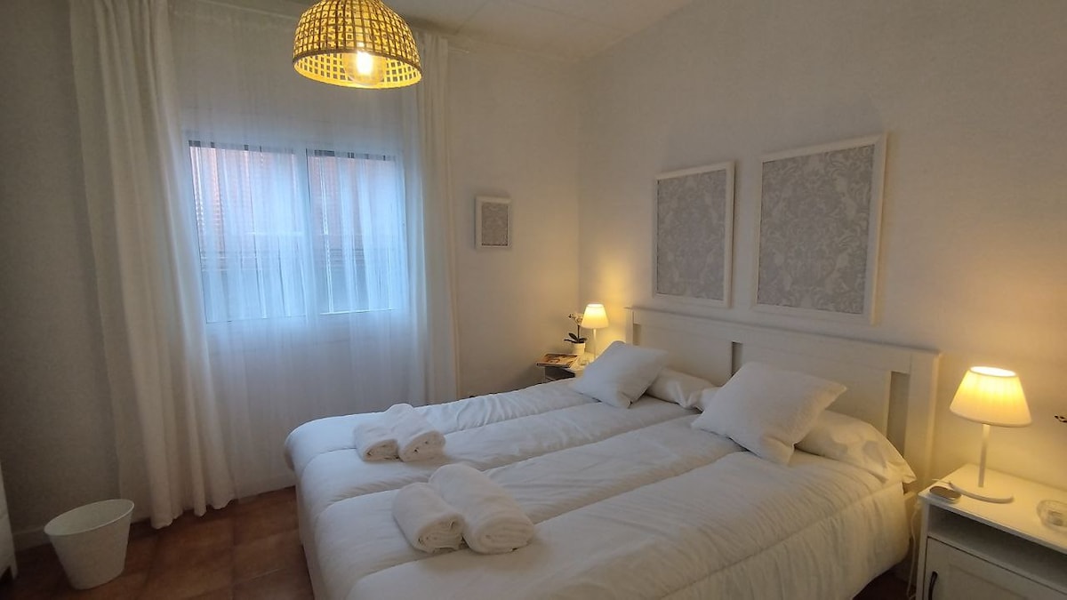 A well-lit bedroom features two spacious beds with white linens and neatly arranged towels. Soft lighting is provided by a bedside lamp, complemented by a decorative pendant light. A window with sheer curtains allows natural light to fill the room, enhancing its bright and airy feel.