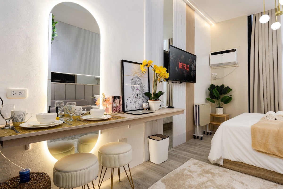 A modern studio space features a streamlined kitchen counter and two stools, elegantly set with tableware. A plush bed is positioned near a wall-mounted TV displaying a Netflix logo. Indoor plants and decorative art enhance the cozy ambiance.