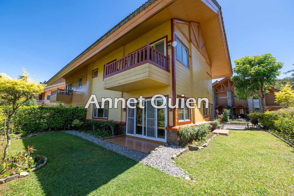 The cabin features a two-story facade with a welcoming balcony above the entrance. A well-maintained garden surrounds the home, highlighted by vibrant greenery and a pebble pathway leading to the door. Large glass doors invite ample natural light into the living space.