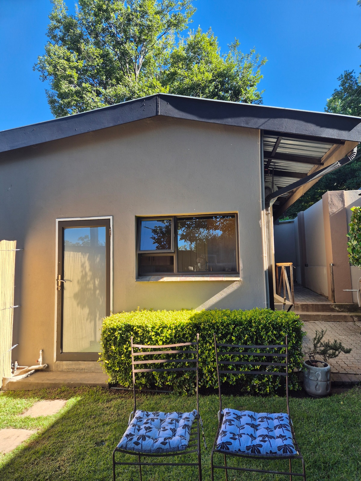 A simple exterior view of a one-story structure is featured, showcasing a large window and a door. Two chairs with patterned cushions are positioned on the well-maintained lawn, bordered by lush greenery. Clear skies provide a bright atmosphere.
