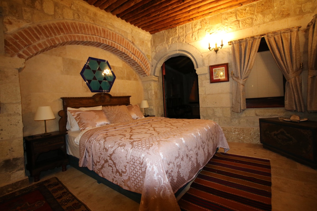 A serene bedroom features a spacious bed adorned with a delicate patterned duvet. Stone walls and wooden beams create a rustic ambiance, complemented by softly lit wall sconces. Traditional textiles and a decorative wall piece add warmth and character to the room.