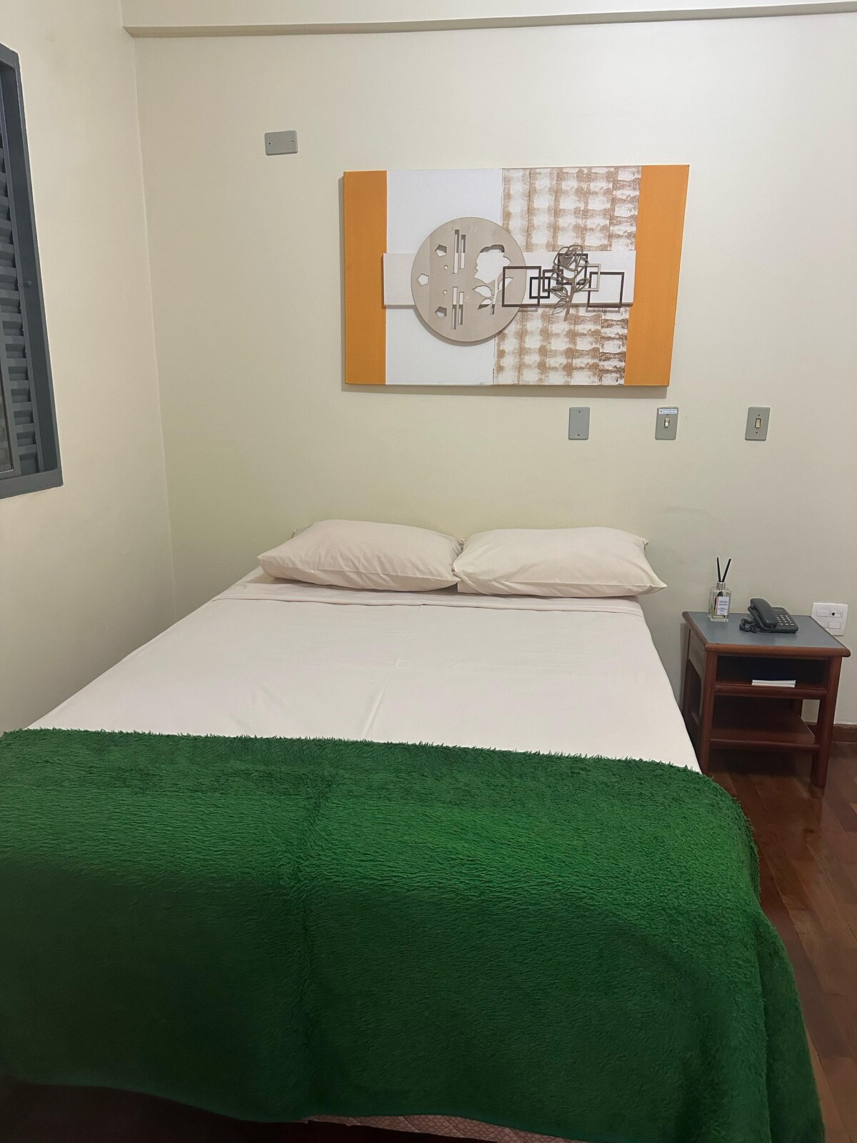 A cozy bedroom features a spacious bed with white linens, complemented by two soft pillows and a green blanket draped across the foot. A piece of abstract wall art adds a modern touch, while a small desk with a phone and a water bottle enhances the utility of the space.