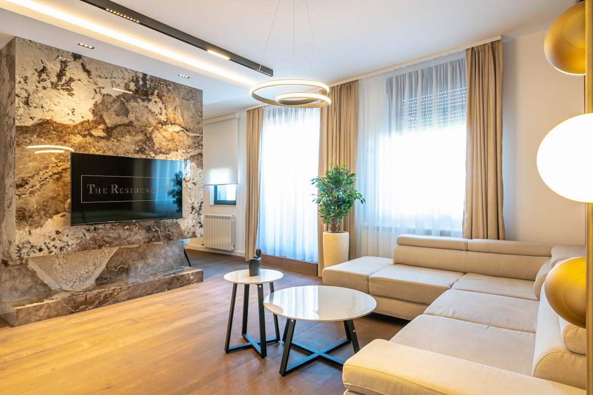 A modern living area features a plush sectional sofa arranged around two circular coffee tables. A striking stone wall with integrated television serves as the focal point. Large windows allow natural light to brighten the space, which is enhanced by tasteful decor and potted greenery.