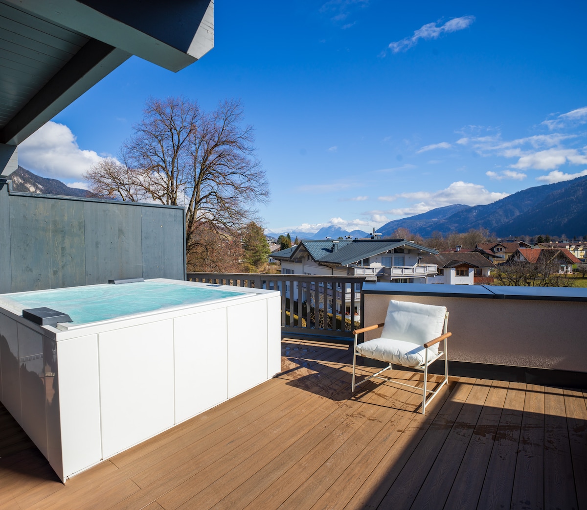 A spacious rooftop terrace showcases a white whirlpool surrounded by wooden decking. A single lounge chair is positioned nearby, offering views of the distant mountains and the clear blue sky, complemented by scattered clouds.