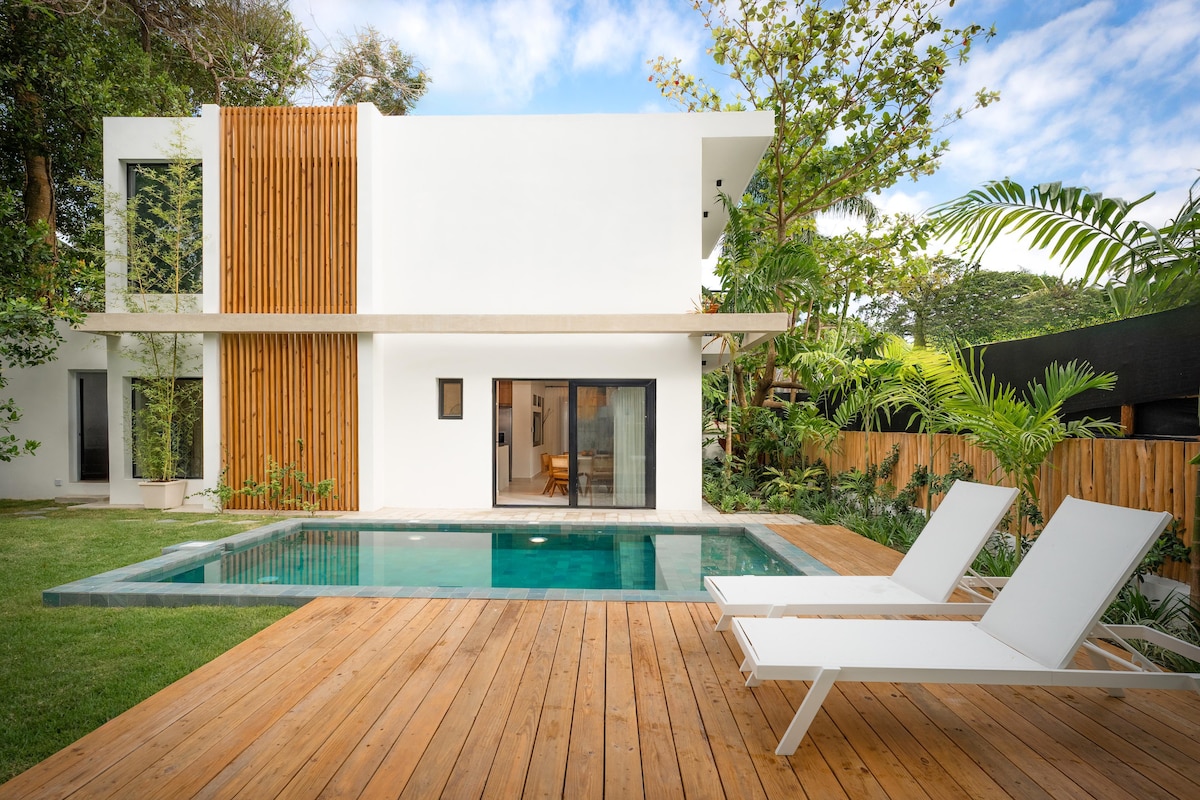 A modern villa is set against a backdrop of lush greenery. The house features a white facade with wooden accents and large windows. A private pool sits in the foreground, surrounded by a wooden deck and tropical plants, creating a serene outdoor space.