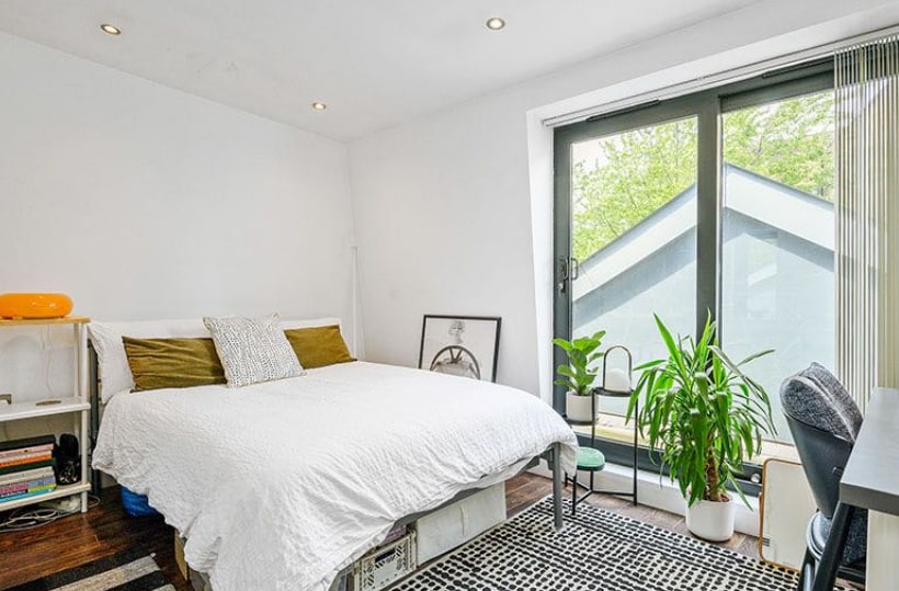 A bright bedroom features a spacious bed with white bedding and decorative pillows. Large glass doors lead to a balcony, allowing natural light to flood the room. A potted plant and a desk with a chair add to the modern decor. A patterned rug completes the space.