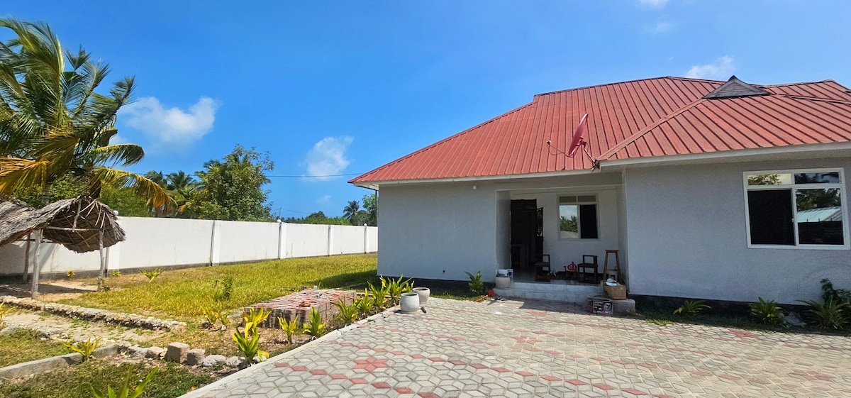 A view of the exterior of a single-story house features a red-roofed structure against a clear blue sky. The well-maintained front yard includes a variety of plants and a paved driveway. A white wall surrounds the property, ensuring privacy.