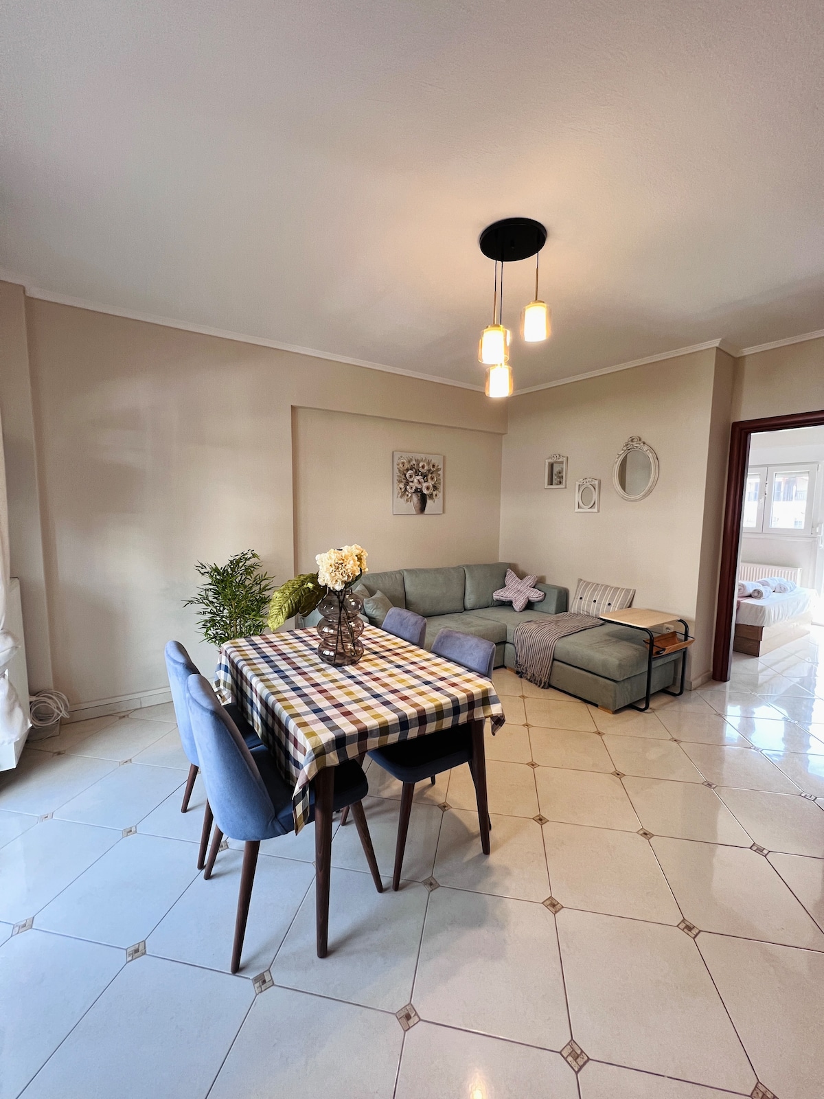 The living and dining area is characterized by a light-coloured tile floor. A wooden dining table with upholstered chairs is set with a decorative vase and a checkered tablecloth, positioned near a cozy lounge area with a sectional sofa.