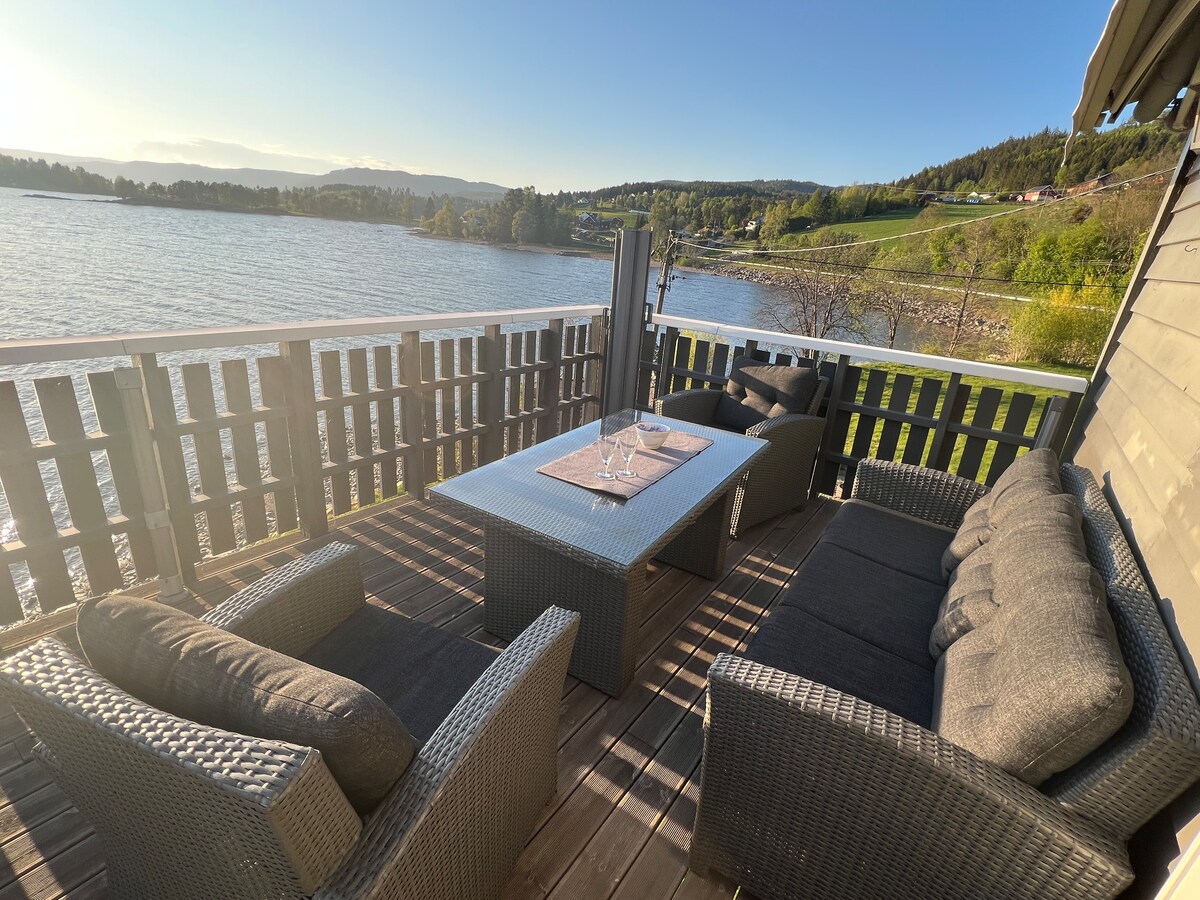 An outdoor seating area is presented on a wooden deck, overlooking Randsfjorden. Comfortable wicker sofas and a table are arranged for relaxation. Sunlight reflects off the water, while scenic views of greenery and hills are visible in the background.