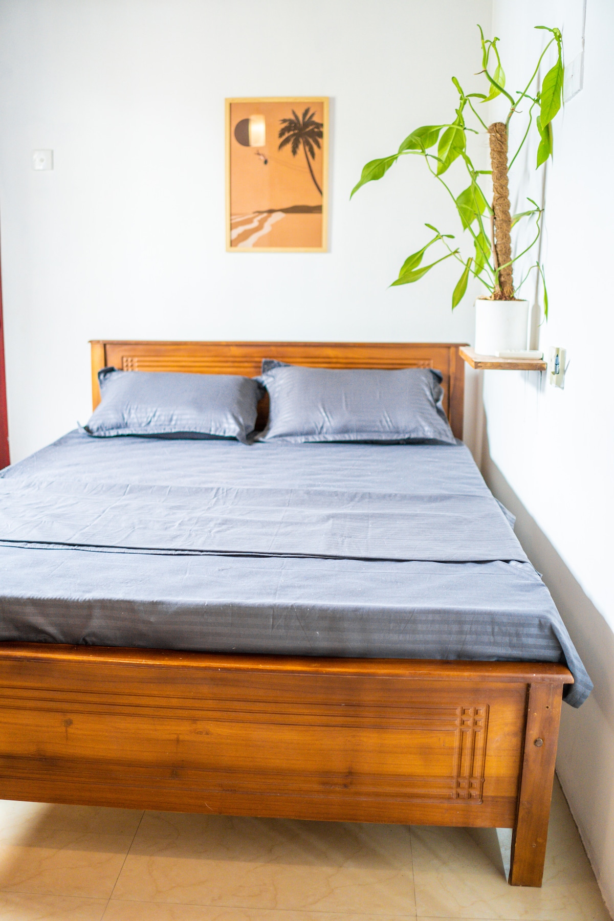 A wooden bed is centered in the room, featuring a simple gray bedding set. Two pillows rest against the headboard. A plant in a white pot is positioned on a wall shelf, and a framed artwork depicting a palm tree is hung above the bed.