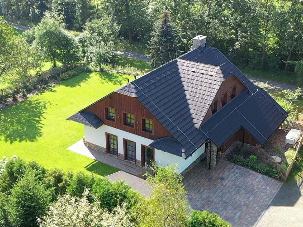 A spacious chalet is depicted from an aerial view, showcasing a sloped roof and a blend of wooden and white exterior. Surrounding green lawns and trees create a natural setting, while stone pathways lead to the entrance.