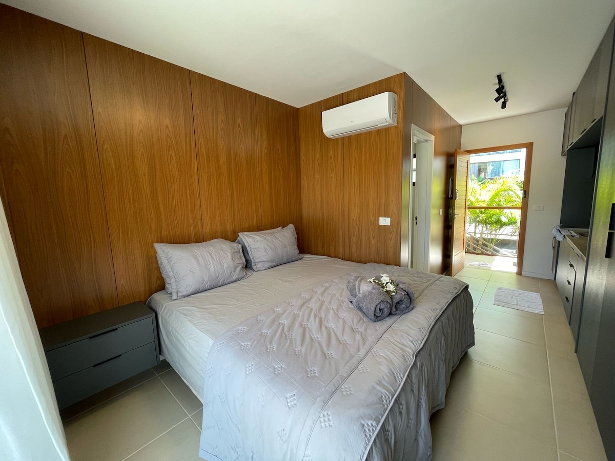 A well-appointed sleeping area features a spacious bed dressed in soft grey bedding. Dark wood paneling adds warmth to the space, complemented by a bedside table. Natural light streams in from a window, which frames views of outdoor greenery, enhancing the inviting atmosphere.