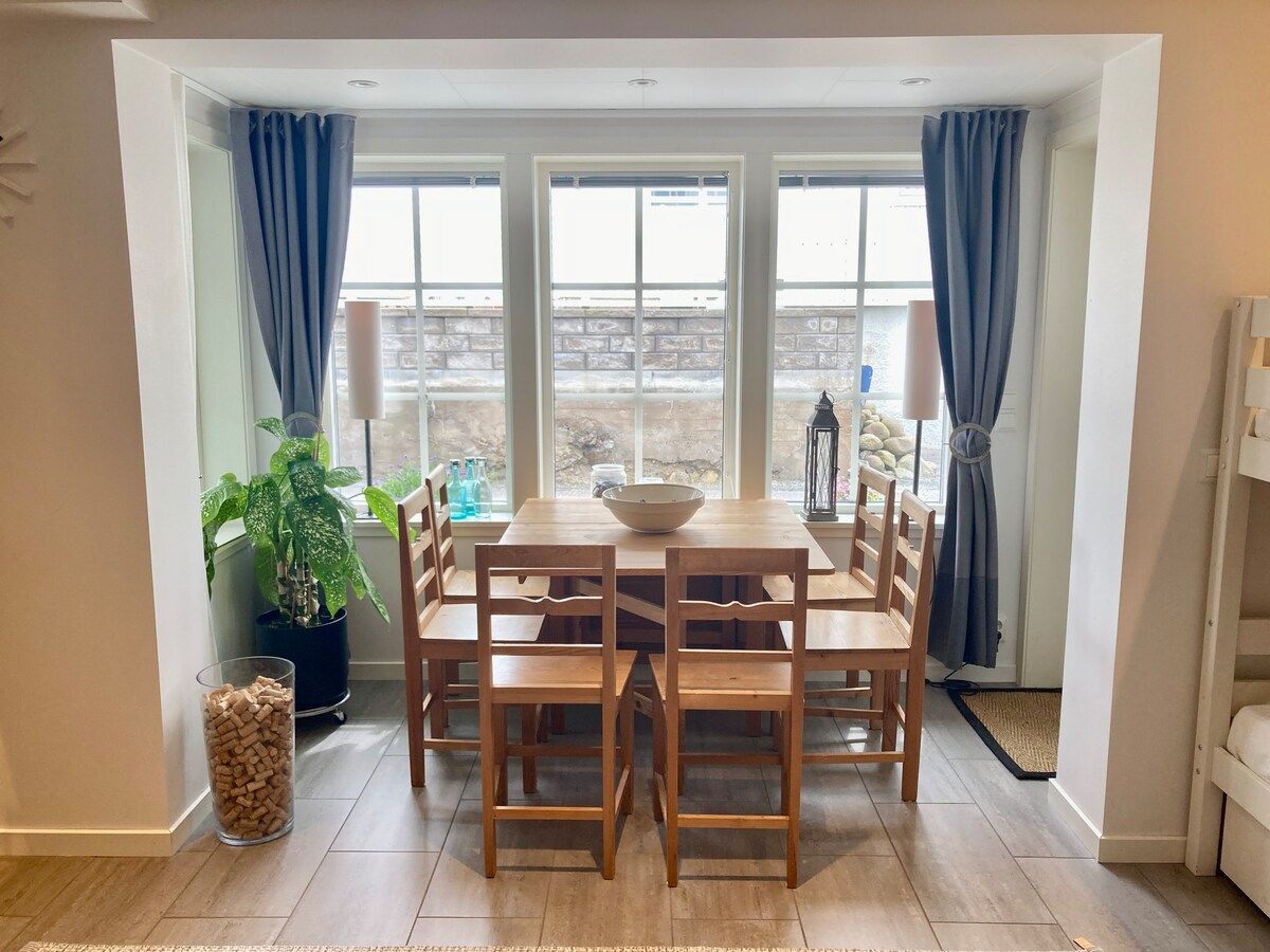 A dining area features a light wooden table surrounded by six wooden chairs. Natural light enters through two large windows, framed by floor-length curtains. A potted plant sits beside the table, and decorative elements are visible on the windowsill.