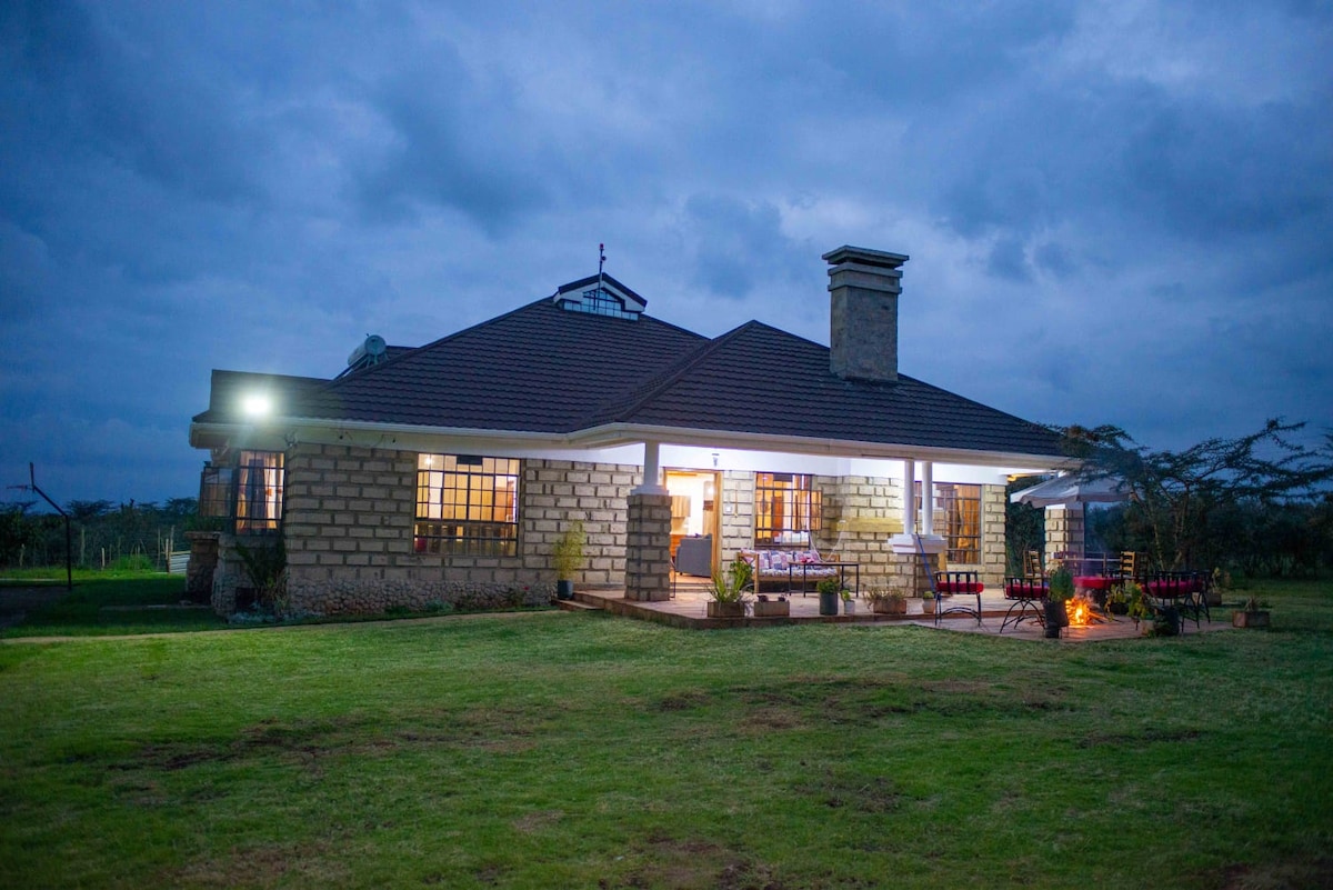 The exterior of a spacious villa is illuminated by soft outdoor lighting during twilight. Large windows allow glimpses of the interior, where a welcoming ambiance is evident. The expansive lawn surrounds the villa, offering a serene view of the natural landscape.