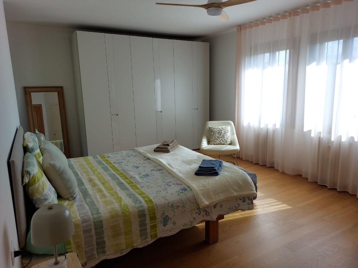 A spacious bedroom is presented with a king-size bed, adorned with a striped blanket and a set of towels. A large wardrobe occupies one side, complemented by an armchair near the window. Natural light fills the room through sheer curtains, enhancing the overall ambiance.