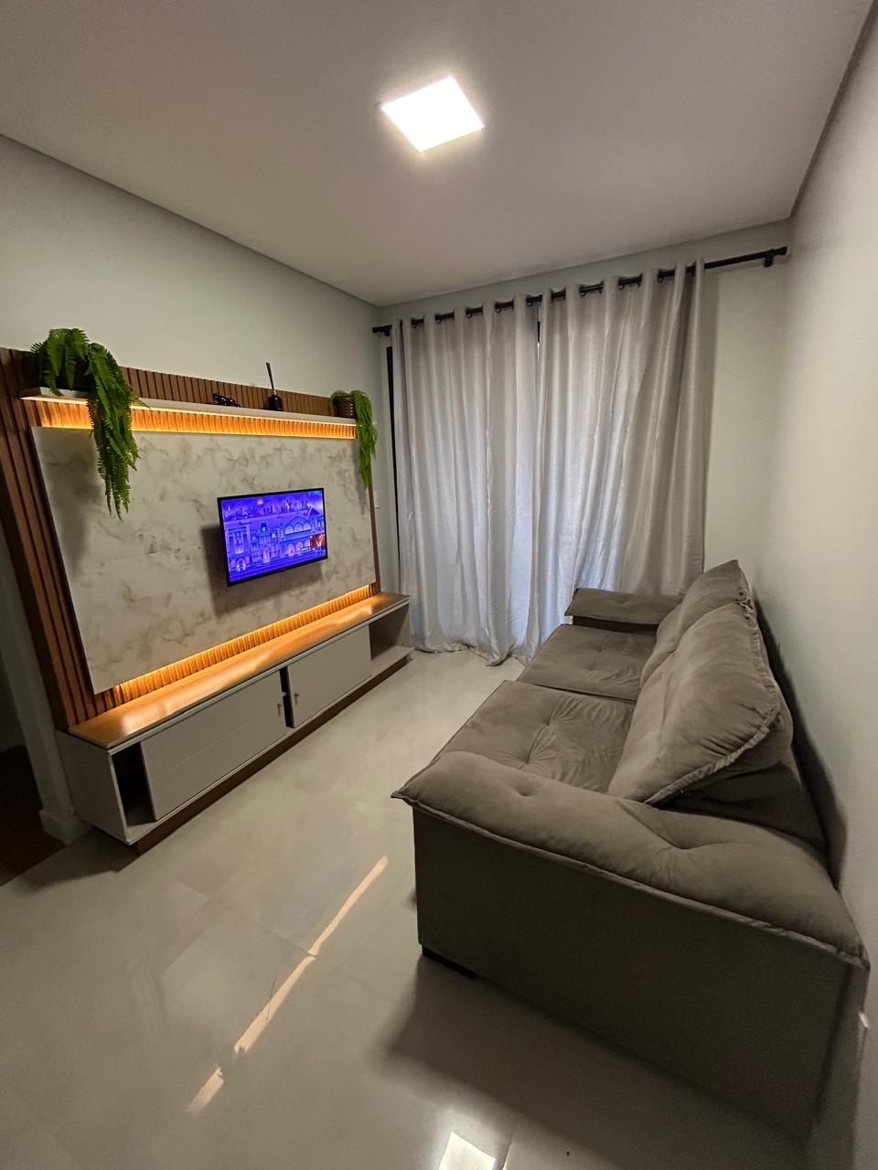 A comfortable seating area is presented with a plush sofa set facing a mounted television. Warm lighting accents the wall behind the TV, which is framed by a marble backdrop. Light-colored curtains are drawn to filter natural light, enhancing the room's brightness.