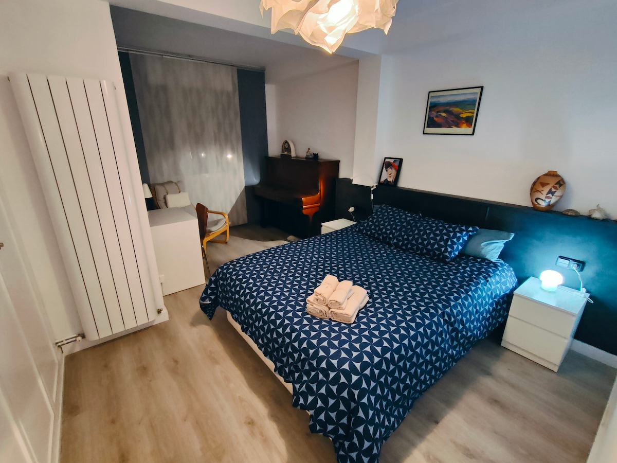 A comfortable bedroom is presented with a spacious bed covered in a patterned blue duvet. Bedside tables with lamps are positioned on either side. A window allows natural light to enter, and a piano is visible in the background, contributing to the room's character.