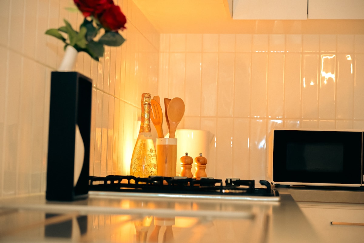 A modern kitchen is showcased with sleek white tiles and warm lighting. A gas stove is visible in the foreground, complemented by a small vase of flowers. Wooden cooking utensils and salt and pepper shakers are neatly arranged on the counter beside a compact microwave.