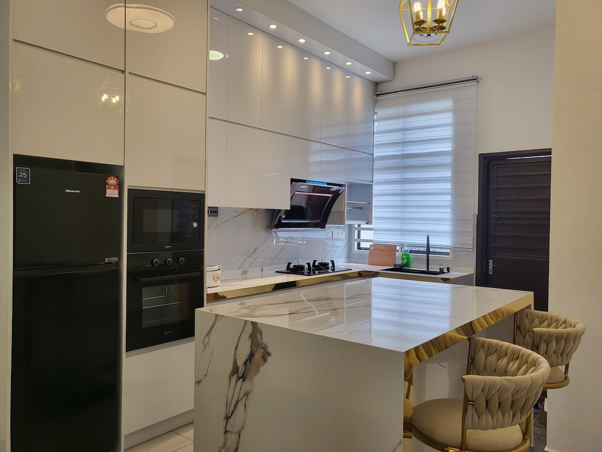 A modern kitchen showcases sleek, glossy cabinetry with an integrated oven and refrigerator. A marble kitchen island with golden accents complements the space. Natural light enters through a nearby window, and a textured, neutral-colored stool is positioned at the island.