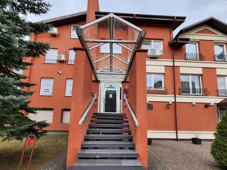 The entrance of the building is illustrated, featuring a set of grey steps leading to a glass-covered portico. The exterior walls are painted in warm orange tones, with windows that are framed elegantly. Lush greenery flanks the entrance, enhancing the welcoming appearance.