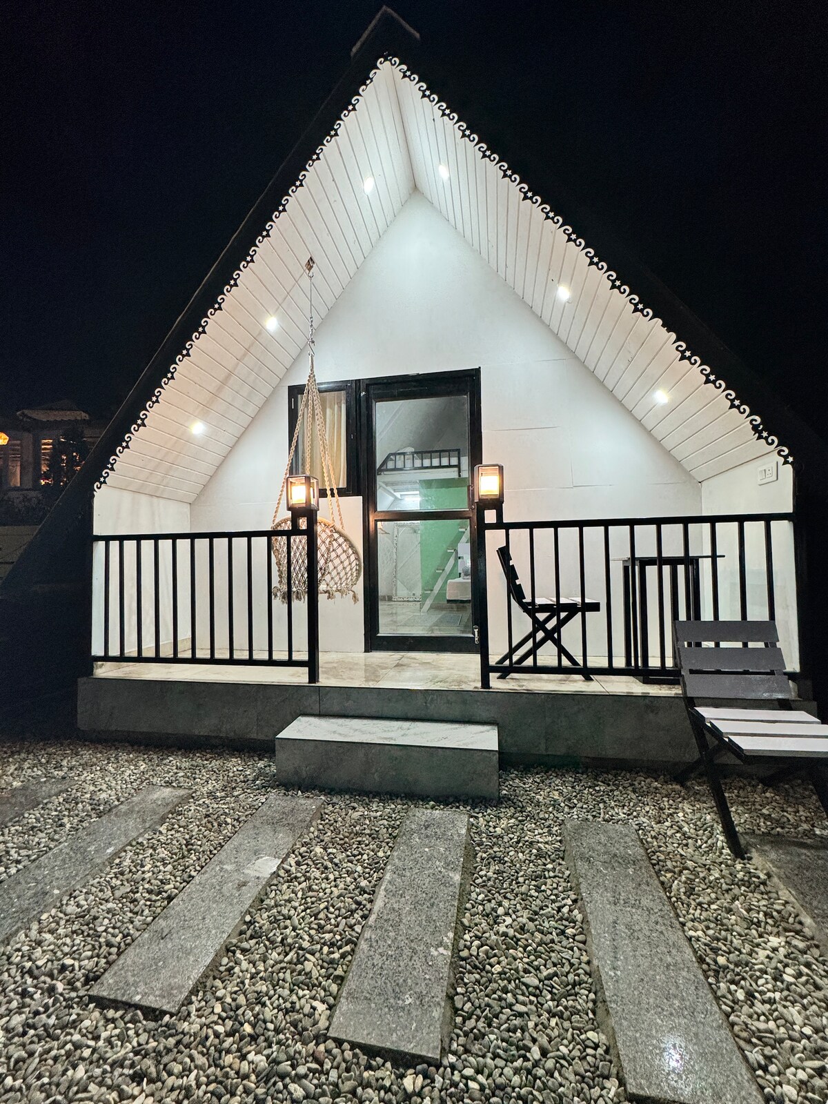 An A-frame structure is illuminated at night, showcasing clean architectural lines. Soft lighting highlights the entrance, where a balcony with a railing is visible. A rocking chair and a striped lounge chair are positioned nearby on a gravel path, leading towards the door.