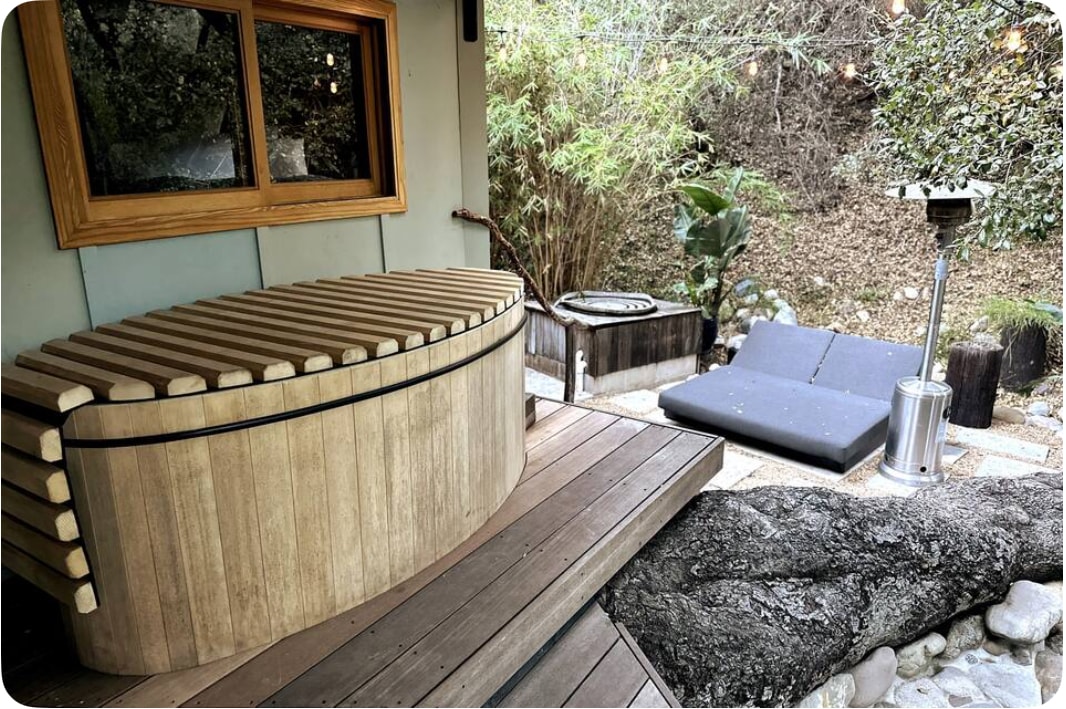 A Canadian cedar jacuzzi is positioned on a wooden deck, surrounded by a natural landscape of bamboo and foliage. Nearby, a seating area features a simple gray cushion on a stone pathway, complemented by a standing heater providing warmth. Soft ambient lighting enhances the serene environment.