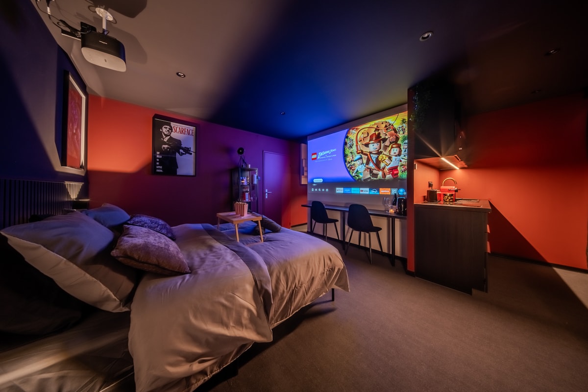 A studio room features a spacious bed arranged with soft pillows. A large screen is mounted on the wall, accompanied by a projector. The space is illuminated in warm tones, with a cozy seating area and a kitchenette visible in the background.