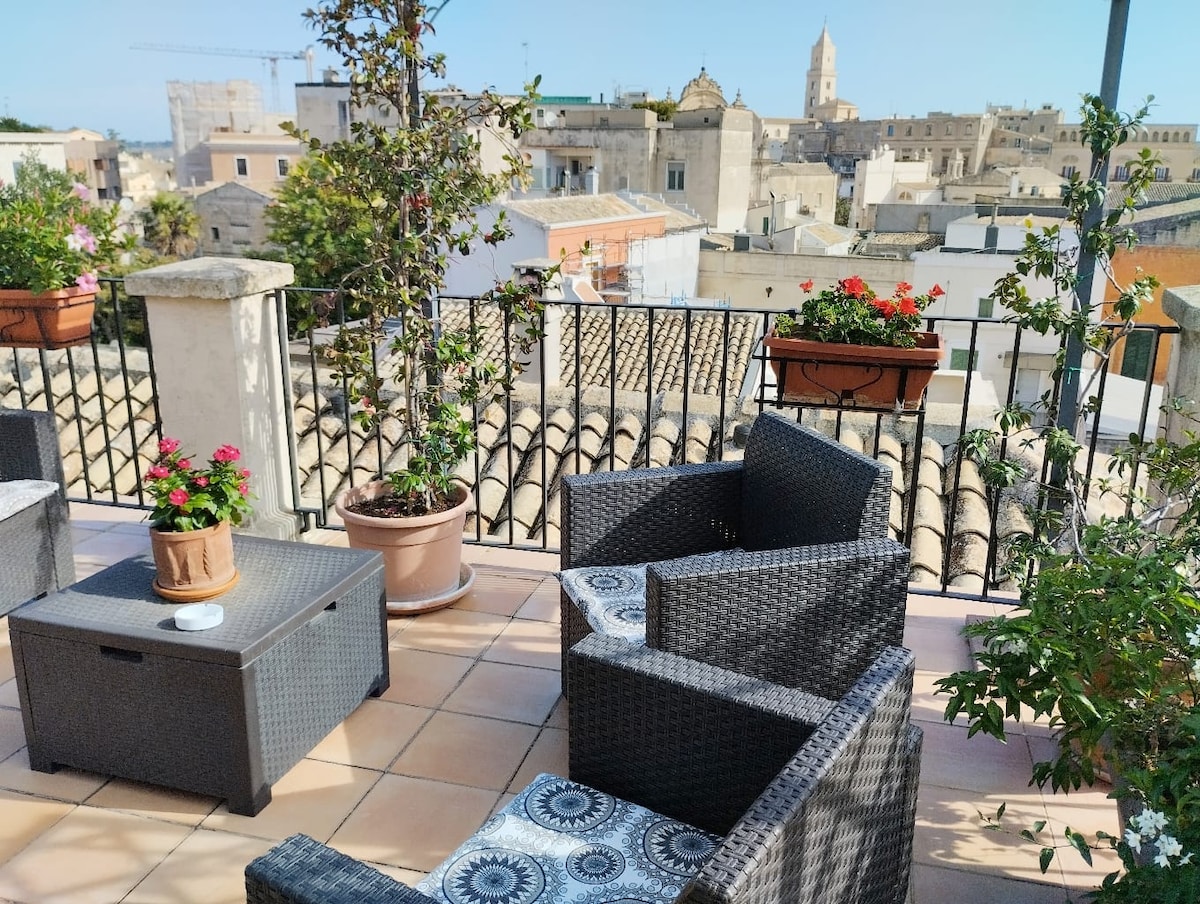 A spacious terrace is furnished with a set of woven chairs and a low table, surrounded by flowering plants in pots. The view includes rooftops and a distant church tower, highlighting the charm of the historic neighborhood.