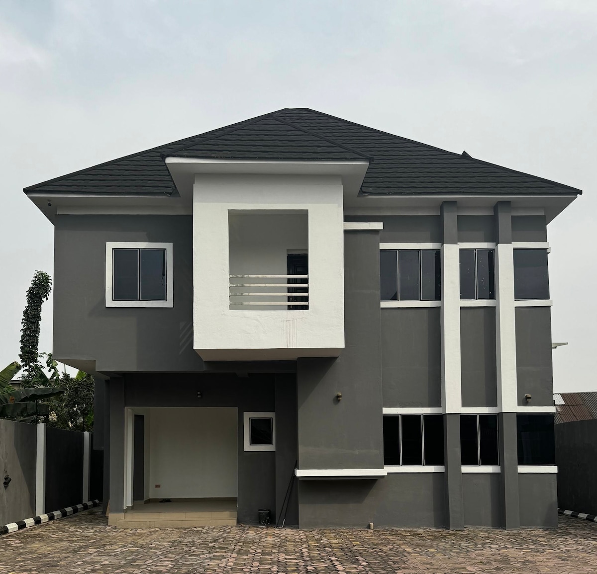 The exterior of a two-story house is presented, featuring a mix of dark grey and white paint. Large windows allow natural light to fill the interior. A covered entryway leads to a spacious front area paved with patterned bricks.