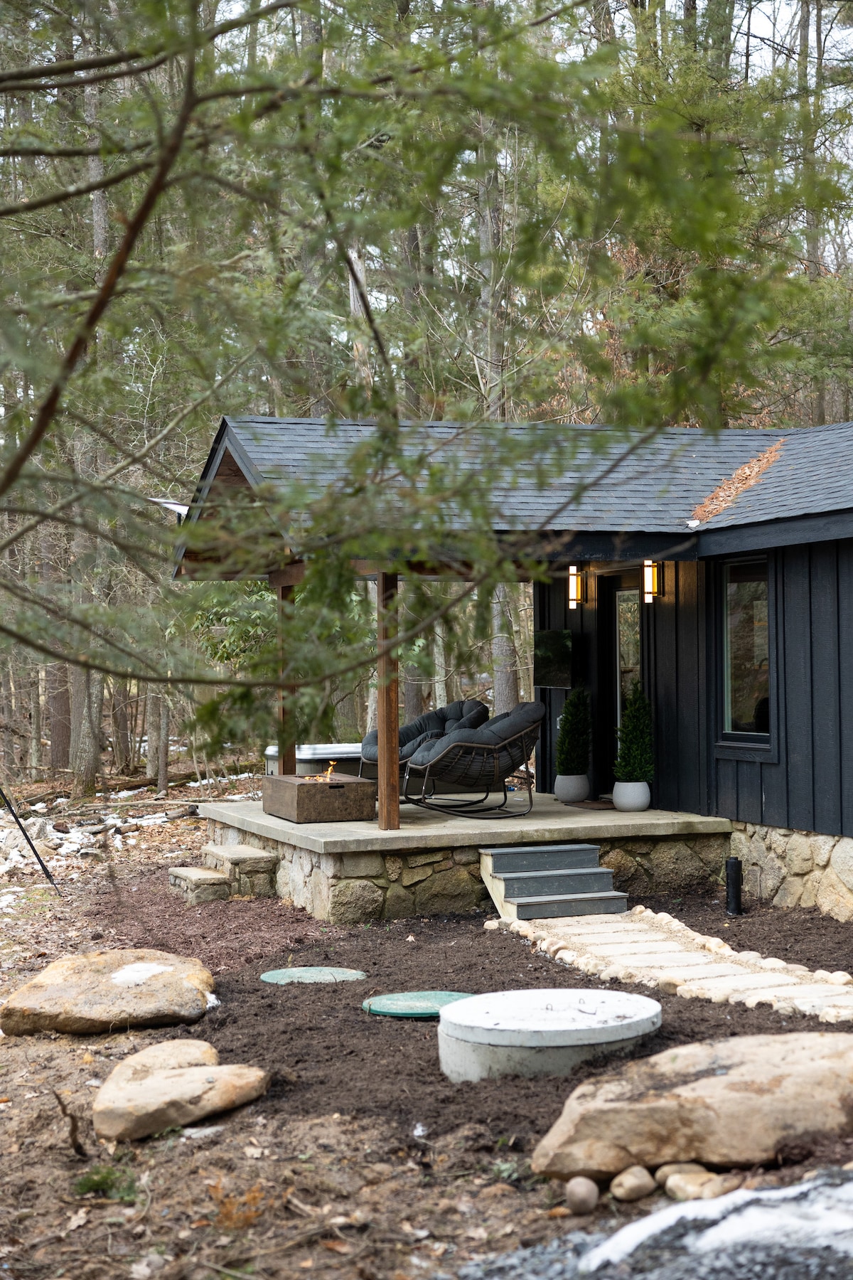 The exterior of the cabin is showcased, framed by trees. A stone patio leads to the entrance, featuring two cozy rocking chairs set beneath a covered area. Natural landscaping elements, including boulders and soil, add to the serene atmosphere.