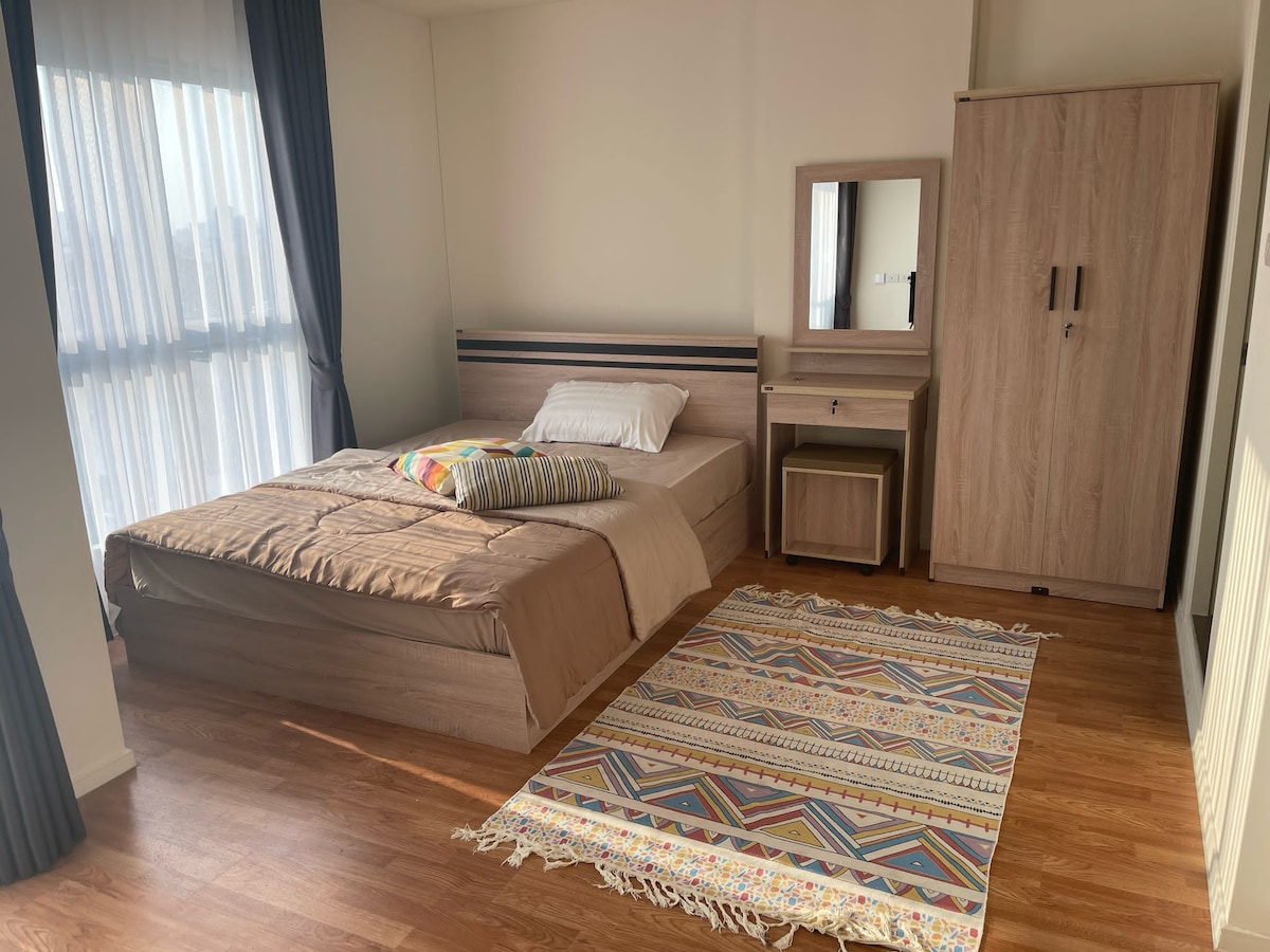 A well-designed studio features a spacious bed with neutral bedding, complemented by a colorful throw pillow. Natural light fills the room through large windows, and a small wooden desk with a mirror adds functionality. A patterned rug enhances the warm wooden flooring.