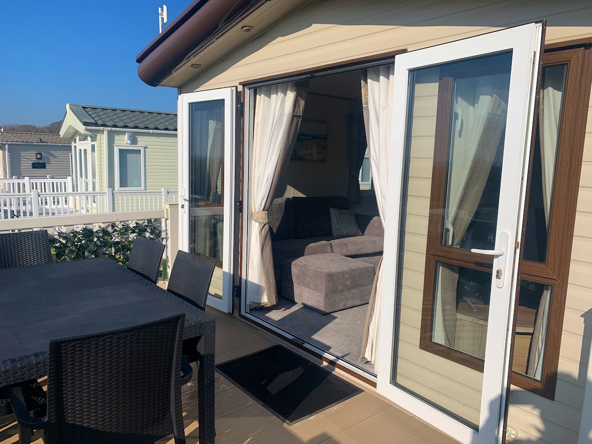 A spacious outdoor deck is featured, furnished with a dining table surrounded by black wicker chairs. The entrance to the caravan is highlighted by large glass doors, revealing a comfortable living area with a gray sectional sofa that invites relaxation.