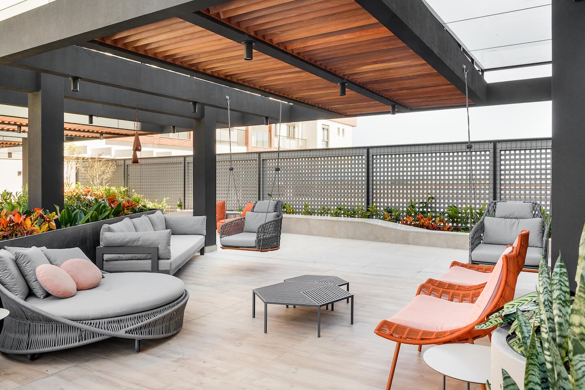 An outdoor lounge area is presented, featuring a mix of modern seating options including circular and angular chairs. A textured gray floor complements the wood-paneled ceiling. Vibrant greenery and decorative plants accentuate the space, providing a calming environment for relaxation.