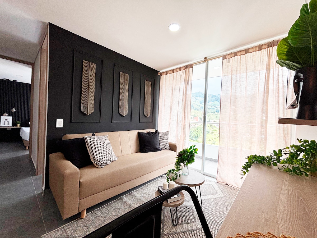 A bright and spacious living area is featured, showcasing a neutral-toned sofa adorned with decorative cushions. Light streams through large glass doors covered with sheer curtains, allowing for a view of the outdoors. Simple greenery is placed on a nearby table, enhancing the serene ambiance.