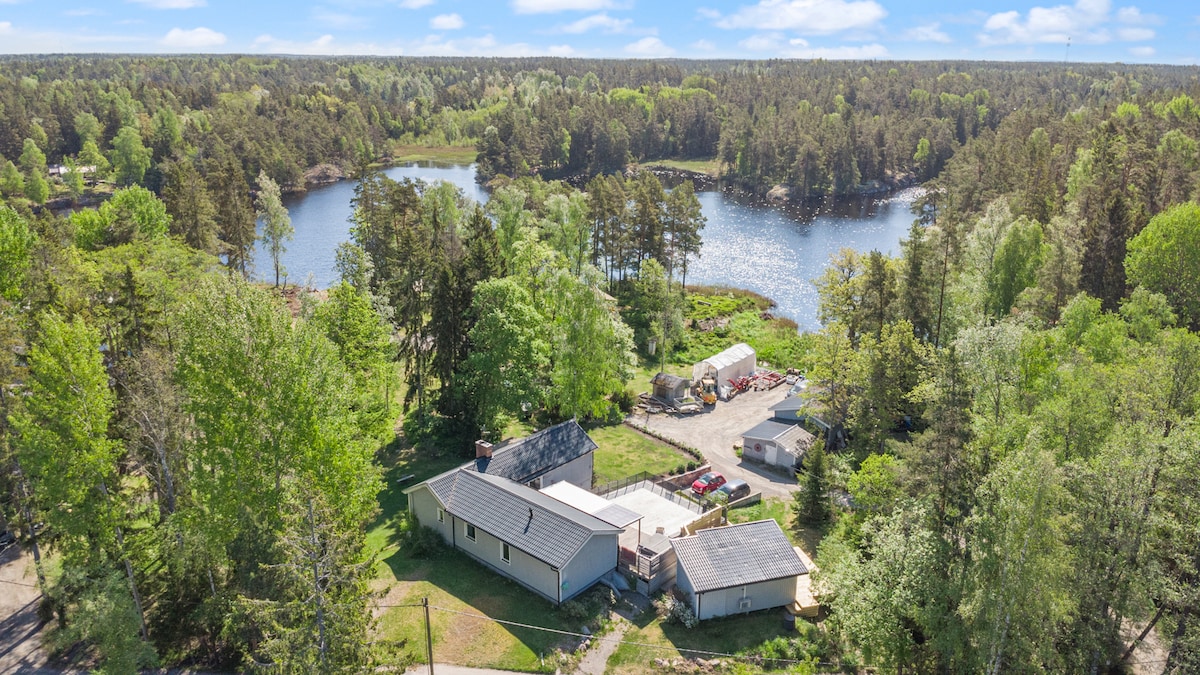 Aerial view of a serene lakeside property surrounded by lush greenery. The water's surface reflects the surrounding trees, while various structures are visible on the property, including a house and additional buildings. A pathway leads through the landscape, offering access to nature.
