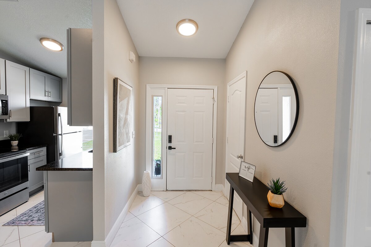 An entryway is highlighted by a round mirror above a sleek, dark console table. Neutral-colored walls and modern lighting fixtures create a bright atmosphere. A welcoming view of the front door is complemented by decorative accents, enhancing the inviting space.