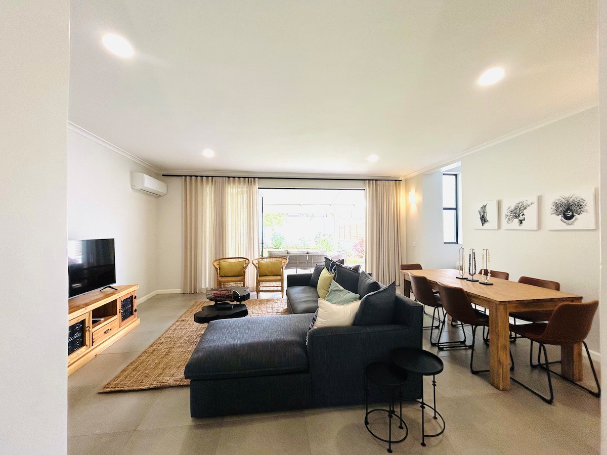 The open living space features a sectional sofa with neutral cushions, accompanied by a wooden dining table surrounded by modern chairs. A large window allows natural light, while decorative wall art is visible in the background. A cozy rug adds warmth to the space.
