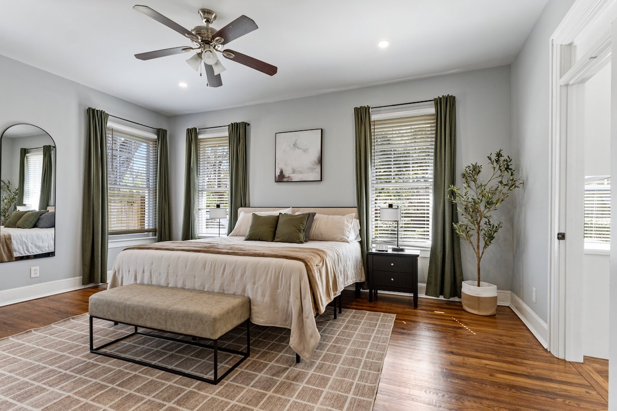 A spacious bedroom features a comfortable bed with neutral linens, accompanied by a decorative bench at the foot. Natural light is allowed in through multiple windows, with green curtains framing the views. A small nightstand and a plant add to the inviting decor.