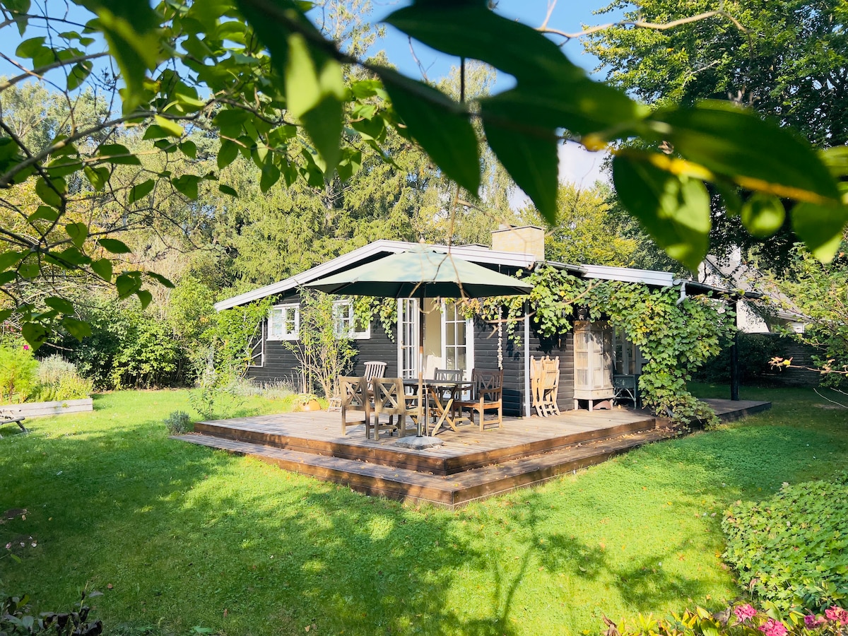 A classic summer house is highlighted by its dark exterior, surrounded by a lush green garden. A patio area features a dining set beneath a sunshade, with ample space for relaxation. Rich foliage frames the property, enhancing the serene outdoor setting.