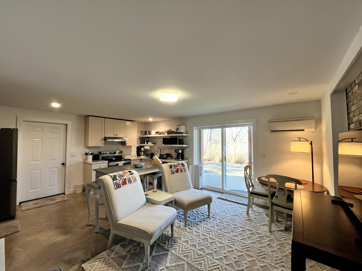 A well-lit sitting area features two chairs with colorful accent pillows and a wood table, creating an inviting space. A full kitchen is visible, along with a sliding glass door that leads to a natural outdoor view. Soft lighting enhances the cozy ambiance.