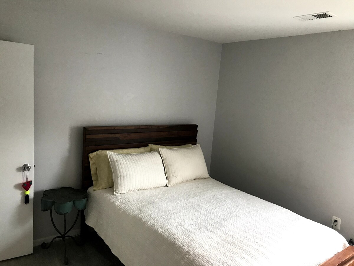 A serene bedroom is furnished with a spacious bed topped with light-colored linens and two crisp pillows. A green side table positioned beside the bed offers a subtle touch. Soft grey walls provide a calming backdrop to the inviting space.