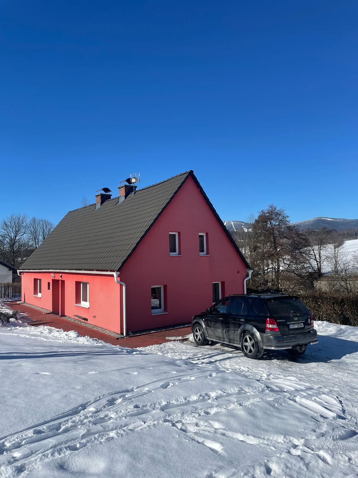 A modern pink house with a sloped roof is set against a clear blue sky. A black vehicle is parked on a driveway covered in snow, while the surrounding landscape features patches of white snow and bare trees.